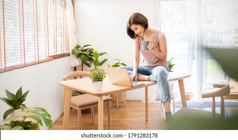 Portrait Young Beautiful Asian Creative Woman Relax Sit On Desk Use Computer Laptop. Smile Happy Asian Girl Drink Coffee At Table Home. Freelance Small Business Asian Owner Sme Work From Home Concept 