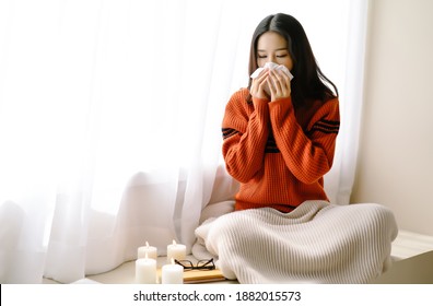 Portrait Of Young Beautiful Asian Woman Sneezing And Blowing Nose With A Tissue Sitting Home In The Chair By The Window. Young Asian Woman Getting Sick With Flu In A Winter Day. Woman With A Cold.