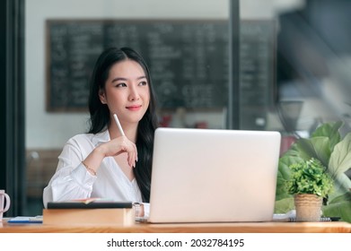 Portrait Of Young Beautiful Asian Woman Holding Stylus Pen, Thinking And Looking Outside While Sitting At Office Desk.