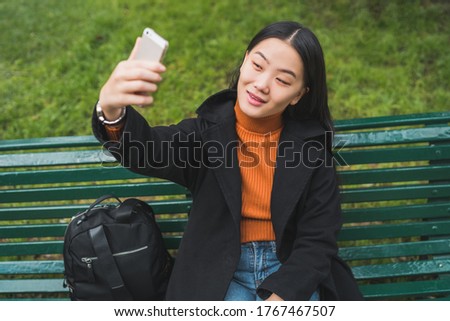 Portrait of young beautiful Asian woman taking a selfie with her mobile phone while sitting on a bench in park.