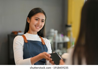 Portrait of young beautiful barista with apron holding over smartphone to help customer pay for purchase a coffee