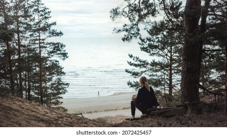 Portrait of a Young Beautiful Blond Woman in a Romantic Nature Atmosphere. Girl is Dressed in Black and is Sitting Alone in a Forest. She is Looking at a Sea Landscape. Man Running in the Distance.