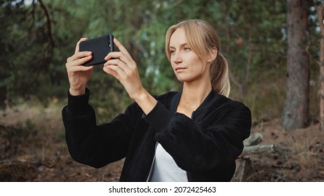 Portrait of a Young Beautiful Blond Woman in a Romantic Nature Atmosphere. Girl is Dressed in Black and is Taking a Picture on Her Mobile Phone. She Sits in a Pine Forest.