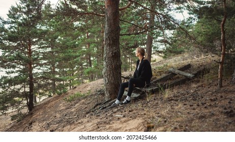Portrait of a Young Beautiful Blond Woman in a Romantic Nature Atmosphere. Girl is Dressed in Black and is Sitting Alone in a Forest. She is Looking at a Sea Landscape.