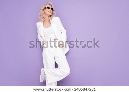 Portrait of young beautiful blond woman wearing nice trendy white suit. Fashion model posing in studio. Fashionable female isolated on violet background. Cheerful and happy. In sunglasses