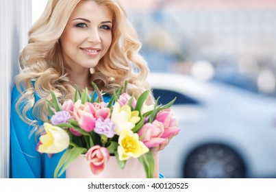 portrait of young beautiful blonde woman with box of flowers by car. flowers in box. spring time outdoor photo. bouquet of flowers in gift box. birthday, March 8, Valentine's Day, romantic