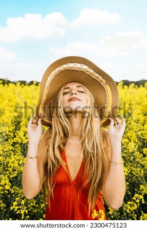 Portrait of a young beautiful blonde woman, wearing a red dress and a hat, amidst a field of blooming yellow rapeseed flowers