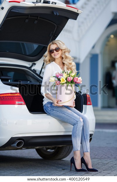 portrait of\
young beautiful blonde woman with box of flowers by car. flowers in\
box. spring time outdoor photo. bouquet of flowers in gift box.\
birthday, March 8, Valentine\'s Day,\
romantic