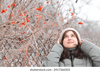 Portrait of young beautiful brunette woman in winter clothes walking on nature, girl standing near viburnum bush and looking up