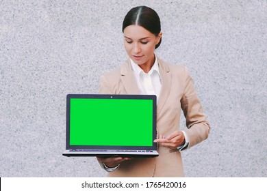 Portrait young beautiful business woman in suit look at empty green screen with copy space on laptop on gray background. Attractive happy girl entrepreneur hold computer with green chroma key in hands
