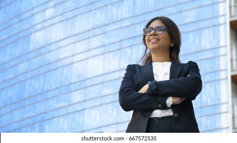 Portrait of young beautiful business woman (student) in suit, glasses, smiling, successful looking at sides, with skyscraper background. Concept: new business, communication, Arab, banker, manager.