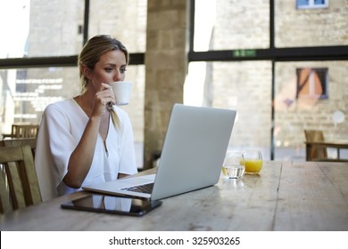 Portrait of a young beautiful businesswomen enjoying coffee during work on portable laptop computer, charming female student using net-book while sitting in cafe bar interior during morning breakfast