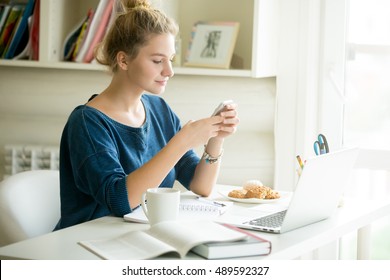 Portrait of young beautiful casual woman holding smartphone, looking at screen, using app or messaging while sitting at modern workplace with laptop, books, coffee and cookies in home office or dorm