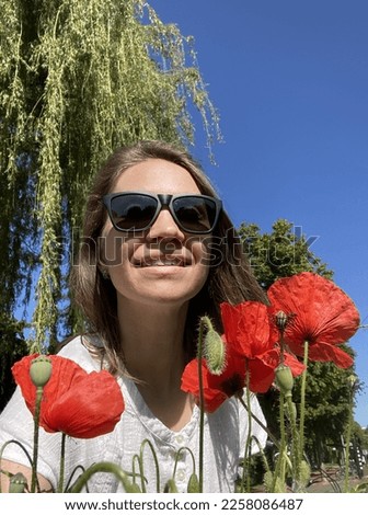portrait of young beautiful European girl in white T-shirt with dark hair in sunglasses, beautiful smile, red poppies in field, background of green trees and blue sky, no clouds in summer on sunny day