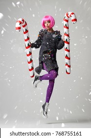 Portrait of young beautiful fashion woman in stylish pink wing and cap hold two big christmas candy cane on grey background under snow