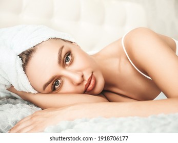 Portrait Of Young Beautiful Female In White Lingerie And Towel On Head. Sexy Carefree Woman Lies On Bed In Posh Apartment Or Hotel Room. Hot Model After Shower