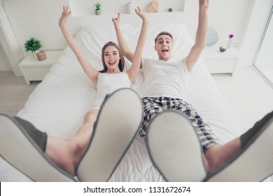 Portrait of young beautiful funky attractive cute foolish couple wearing pajama lying together on bed in apartment, laughing, raising hands and legs up