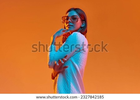 Portrait of young beautiful girl in casual clothes and trendy sunglasses posing against orange studio background in neon light. Concept of human emotions, youth, feelings, fashion, lifestyle, ad