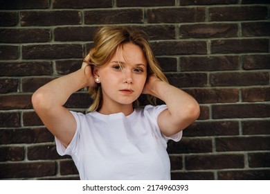 Portrait Of A Young Beautiful Girl Looking Into The Camera Against A Brick Wall. A Fashionable Teenager In Casual Summer Clothes. Her Hair Is Blown Away By The Wind And Falls On Her Face.