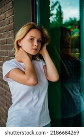 Portrait Of A Young Beautiful Girl Looking Into The Camera Against A Brick Wall. A Fashionable Teenager In Casual Summer Clothes. Her Hair Is Blown Away By The Wind And Falls On Her Face.