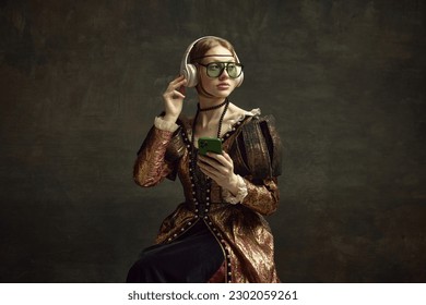 Portrait of young beautiful girl in vintage dress, trendy sunglasses, headphones and mobile phone against dark green background. Concept of history, renaissance art, comparison of eras, social media