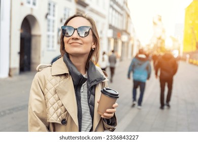 Portrait young beautiful happy smiling woman drinking coffee to go tea in eco sustainable paper cup wear biege trench coat enjoy walking european city street. Stylish female person urban lifestyle