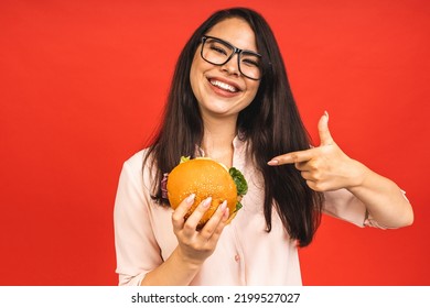 Portrait Of Young Beautiful Hungry Woman Eating Burger. Isolated Portrait Of Student With Fast Food Over Red Background. Diet Concept.