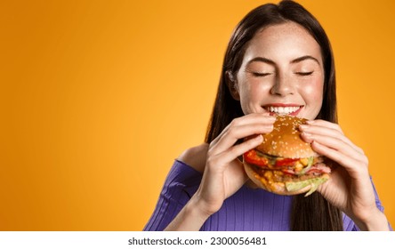 Portrait of young beautiful hungry woman eating burger. Isolated portrait of student with fast food over orange background. Diet concept. - Powered by Shutterstock