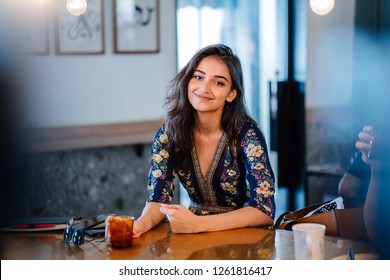Portrait of a young and beautiful Indian woman sitting at a counter in a stylish coffee shop and enjoying a drink on the weekend. She has her notebook and a pair of sunglasses next to her. 