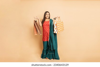 Portrait of young beautiful indian woman wearing traditional outfit holding shopping bags standing isolated on beige studio background. Diwali celebration and festive sale concept.Full length.