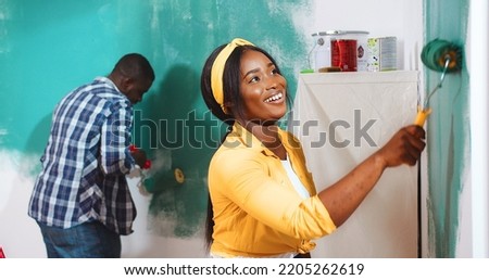 Portrait of young beautiful joyful smiling African American wife painting wall with her husband in green color with paint roller brush, redesigning room Side view home renovation apartment improvement
