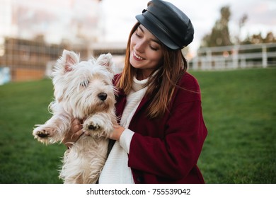 Portrait of young beautiful lady in black cap and coat holding in hands her small cute dog while spending time in park