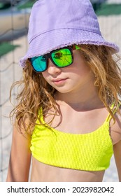 Portrait of a young beautiful model girl with hat and sunglasses posing on the beach. Wearing a bright yellow bikini swimsuit. Summer day, white sand. High quality photo