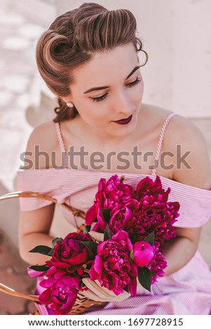 Portrait young beautiful retro style woman in pink stripped dress and pin up hairstyle sit in park with basket of flowers. Attractive tender stylish vintage girl posing with peonies flowers outdoors