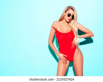 Portrait of young beautiful sexy woman with large breasts. Carefree model wearing pure red lingerie. Hot tanned blonde posing near blue wall in studio in summer swimwear bathing suit. In sunglasses