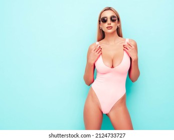 Portrait of young beautiful sexy woman. Carefree model wearing pure pink lingerie with big breasts. Hot tanned blonde posing near blue wall in studio in summer swimwear bathing suit. In sunglasses