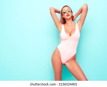 Portrait of young beautiful sexy woman. Carefree model wearing pure pink lingerie with big breasts. Hot tanned blonde posing near blue wall in studio in summer swimwear bathing suit. In sunglasses
