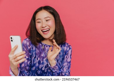 Portrait Of Young Beautiful Smiling Asian Girl With Opened Mouth Holding Phone And Taking Selfie With Raised Hands While Standing Over Isolated Red Background