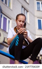 A Portrait Of A Young Beautiful Smiling Brunette Middle School Age Teenager Girl With Long Dark Hair. Urban View. Summer Vacations. A Girl Holds A Smartphone In Her Hands And Looks At The Screen.