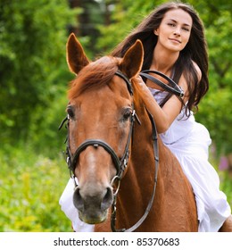 Portrait of young beautiful smiling brunette woman wearing white dress riding dark horse at summer green forest.