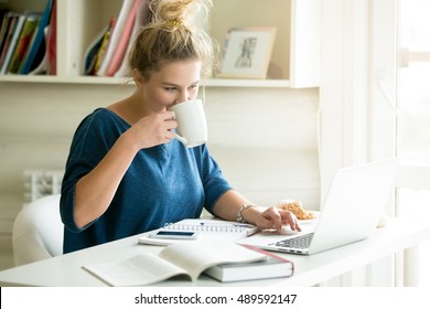 Portrait of young beautiful smiling casual woman sitting at office desk enjoying her cup of coffee while working or studying on laptop computer at small home office or in the student dorm. Indoors