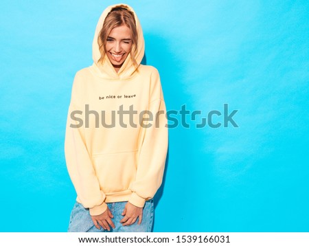 Portrait of young beautiful smiling girl in trendy summer hipster yellow hoodie.Sexy carefree woman posing near blue wall. Positive model having fun.Winks and shows tongue