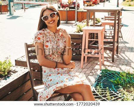 Portrait of young beautiful smiling  girl in trendy summer sundress.Sexy carefree woman sitting in veranda cafe on the street background at sunset. Positive model outdoors in sunglasses