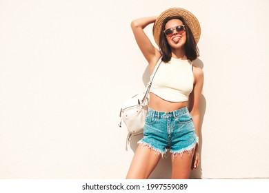 Portrait of young beautiful smiling hipster woman in trendy summer jeans shorts and hat. Sexy carefree model posing in the street near white wall. Positive model outdoors with briefcase.Shows tongue