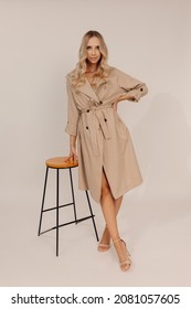 Portrait Of Young Beautiful Smiling Woman With Professional Make-up, Long Blond Wavy Hair Standing Near Chair In Beige Trench Coat Shoes High-heels. Real People. Full Length Photo. Looking At Camera