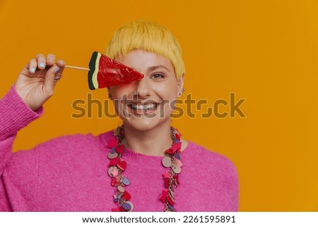 Portrait of young beautiful stylish short-haired smiling woman in necklace covering her eye with a watermelon lollipop and looking at camera while standing over isolated orange background