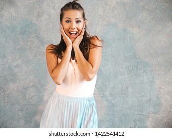 Portrait of Young beautiful surprised woman looking at camera. Trendy girl in casual summer clothes. Shocked female posing near gray wall in studio.Hands near face