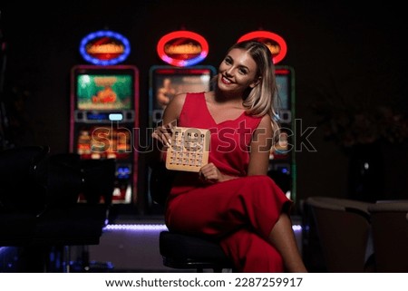Portrait of a Young Beautiful and Well Dressed Caucasian Girl in Red Dress Playing in the Casino and Celebrate Holding Bingo Wood Game Card