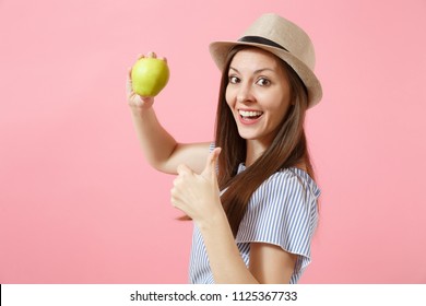 Portrait of young beautiful woman in blue dress, summer straw hat holding, eating green fresh apple fruit isolated on pink background. Healthy lifestyle, people, sincere emotions concept. Copy space