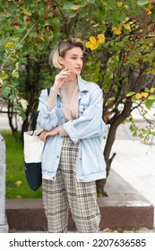 Portrait Of A Young Beautiful Woman In A Denim Jacket Outside, Stands Near A Green Bush